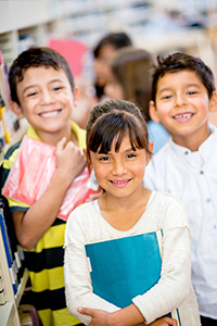 three children smiling, girl in front with brown hair holding blue book, two boys behind her with brown hair