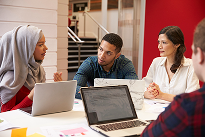 Four teachers sit around a table in a classroom having a discussion. Each teacher has papers and a laptop in front of them. 