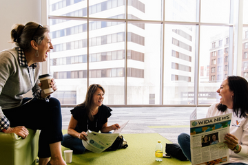 Three people laughing and reading a WIDA News newspaper in front of a large window