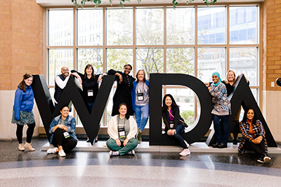 Group of conference attendees standing on and around a large sign spelling out WIDA with the ‘I’ missing. 