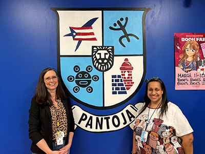 Two people stand on either side of a school crest that’s colorfully painted on a wall.  